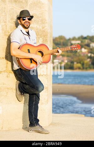 The smiling singing cheerful guitarist is playing guitar outdoors at riverside. Stock Photo