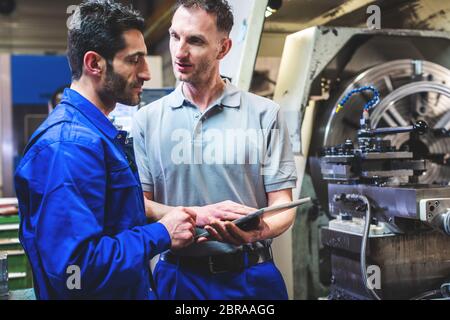 Two workers discussing a project in front of CNC lathe machine writing in clipboard Stock Photo