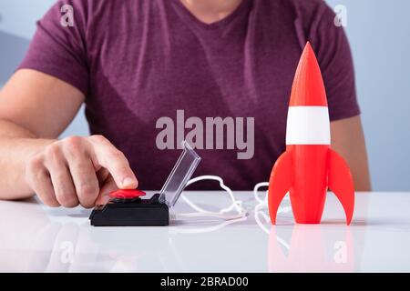 Man's Hand Launching Rocket By Pressing Red Button Over Reflective Desk Stock Photo