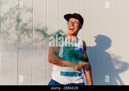 Excited handsome man in eyeglasses waving a smoke bomb, having fun outdoors. Dressed in stylish clothes and cap. Stock Photo