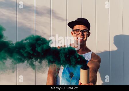 Cheerful attractive young man standing with green smoke flare outdoors, wearing trendy clothes, eyeglasses and rap cap. Stock Photo