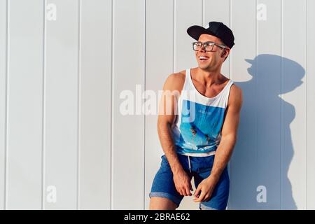 Funny attractive man in eyeglasses and cap posing near the urban wall with copy spac, wearing summer singlet and shorts. Outdoors. Stock Photo