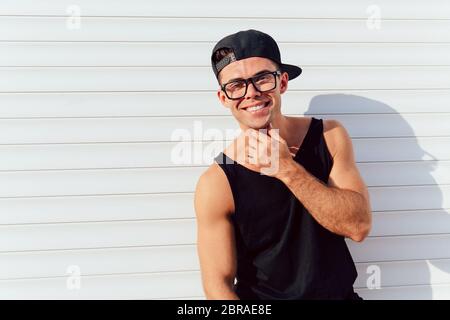 Outdoor photo of stylish young man in fashionable clothes, eyeglasses, rap cap, posing near the white wall, looking at camera. Outdoors. Stock Photo