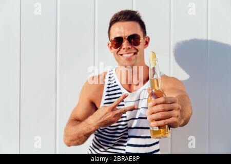 Handsome funny man in sunglasses showing two fingers, peace gesture, while standing with bottle of beverage, outdoors. Dressed in stylish clothes. Stock Photo