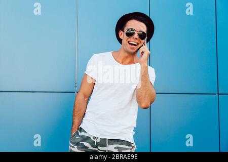 Handsome happy guy in sunglasses and black hat, having pleasant phone conversation, standing outdoors. Dressed in t-shirt and shorts. Stock Photo
