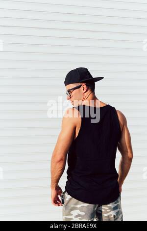 Back view on strong muscular young man standing against the white wall, wearing black singlet, shorts and cap. Outdoors. Stock Photo