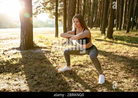 Full length view of attractive sportive woman in tank top and leggings, doing exercises, stretching her body, workout in the park. Sport concept. Stock Photo