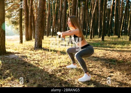 Side view of attractive sportive woman in tank top and leggings, doing squats, stretching body, workout in the wood. Sport concept. Stock Photo