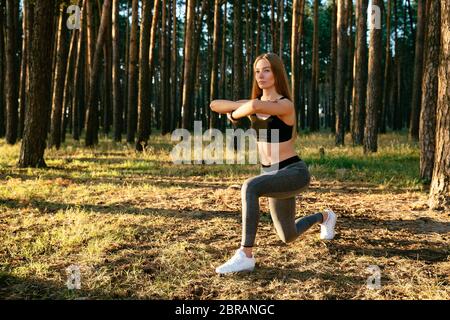 Full length view of young concentrated sportive woman in tank top and leggings, doing exercises, stretching her body, workout in the park. Sport conce Stock Photo