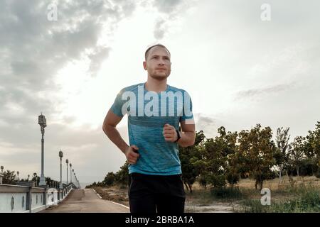 portrait of Caucasian guy in a blue t-shirt and black shorts who trains and runs on the asphalt track during sunset Stock Photo