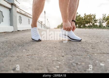 portrait of Caucasian guy in a blue t-shirt and black shorts,who ties his laces on white sports shoes before Jogging Stock Photo