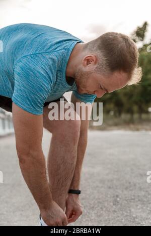 portrait of Caucasian guy in a blue t-shirt and black shorts,who ties his laces on white sports shoes before Jogging Stock Photo