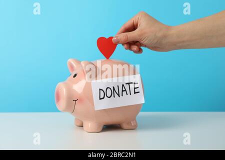 Female hand puts heart in piggy bank with text Donate against blue background Stock Photo