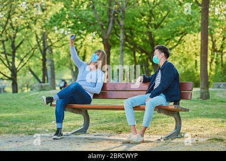 Man and woman wearing a face mask on a park bench in a municipal park, doing selfie, Corona crisis, Regensburg, Bavaria, Germany Stock Photo