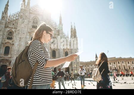 Girl tourist or student with a backpack looks at the map. In the background the Duomo Cathedral in Milan in Italy and unrecognizable people. Stock Photo