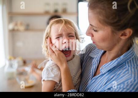 A mother holding a crying toddler daughter indoors in kitchen. Stock Photo