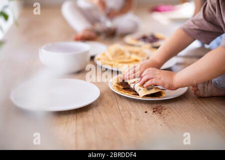 Unrecognizable two small children indoors in kitchen eating pancakes. Stock Photo
