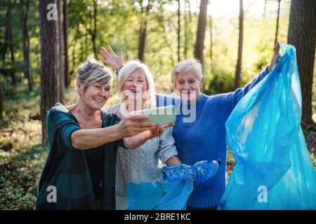 Senior women friends picking up litter outdoors in forest, taking selfie. Stock Photo