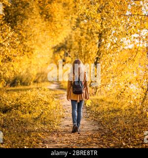 Woman in autumn park, back view. Adult girl walking away alone on path in autumn forest. Lonely young woman with backpack in beige autumn jacket. Beau Stock Photo
