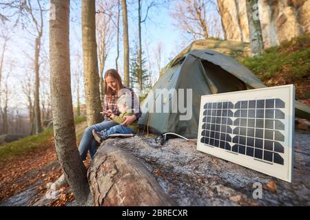 Focus on small solar panel in a campsite on a sunny autumn day against the background of a young family. Mom and child watching something at a mobile phone while enjoying outdoor activities Stock Photo