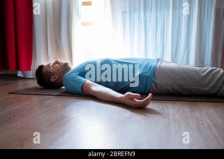 Young man meditating on a floor and lying in Shavasana pose at his living room. Stock Photo