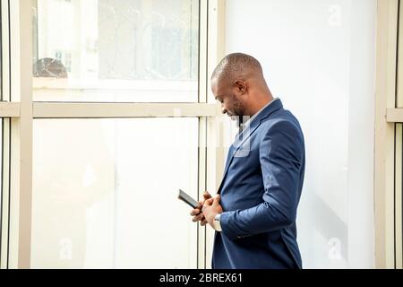 young successful african business man, CEO using his phone standing next to a open glass window in his office Stock Photo