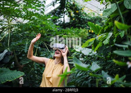 Front view of young woman standing in botanical garden, using VR glasses. Stock Photo