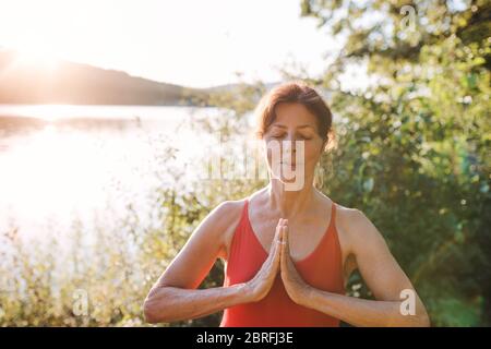 Senior woman in swimsuit standing by lake outdoors doing yoga exercise.
