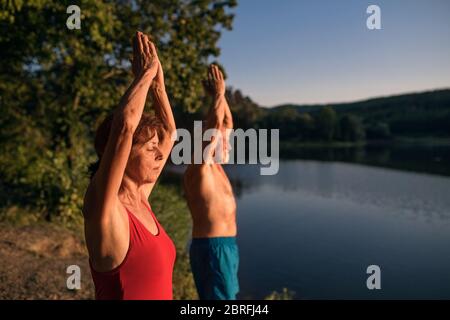 Senior couple in swimsuit standing by lake outdoors doing yoga. Stock Photo