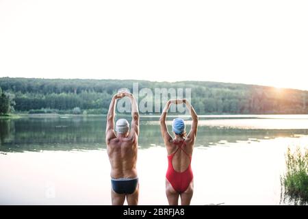 Rear view of senior couple in swimsuit stretching by lake outdoors before swimming. Stock Photo