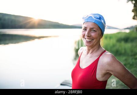 Senior woman in swimsuit standing by lake outdoors before swimming.