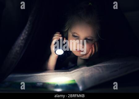 little girl reading book with flashlight under the blanket in dark room Stock Photo