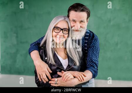 Studio shot of cheerful stylish bearded man embracing his gray haired charming wife from the back and looking at camera with smile, standing on grenn Stock Photo