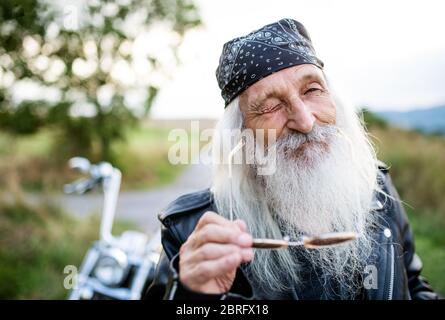 Senior man traveller with motorbike in countryside, winking. Stock Photo