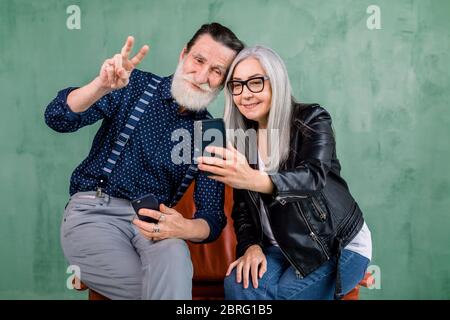 Attractive stylish couple, bearded man and gray haired lady, sitting together on the red chair and using the phone camera for making photo, and Stock Photo