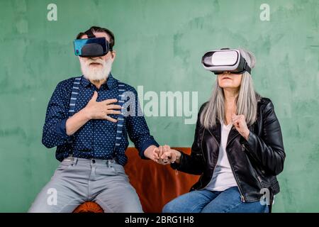 Portrait of surprised excited elderly man and woman, sitting together in red chair near green wall, holding hands, and enjoying the virtual reality Stock Photo