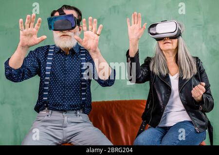 Front view of excited cheerful elderly man and woman, sitting together on red soft chair on green background, and using vr glasses headset, touching Stock Photo