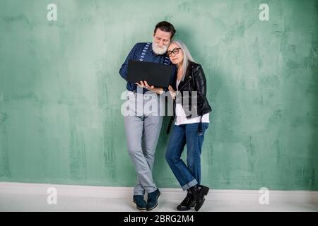 Full length studio portrait of attractive happy modern senior couple, man and woman, wearing trendy stylish clothes, standing together near green wall Stock Photo