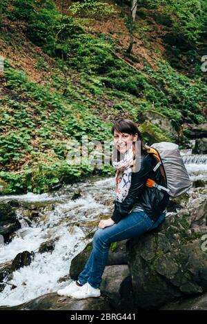 Happy smiling tourist girl in blue jeans, striped shirt and black jacket with backpack near to the cold fresh river at the waterfall Shypit, cascade Stock Photo