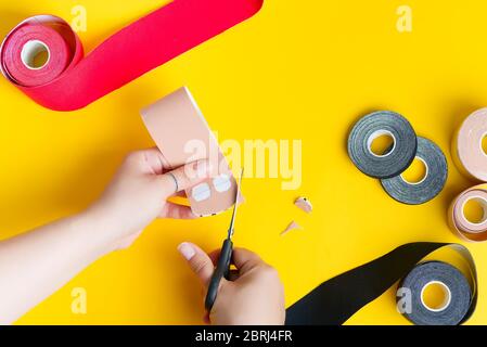 Starting process of Kinesiotaping by womans hands with beige rolling tape and scissors above yellow background, copy space. Step by step. Kinesio trea Stock Photo