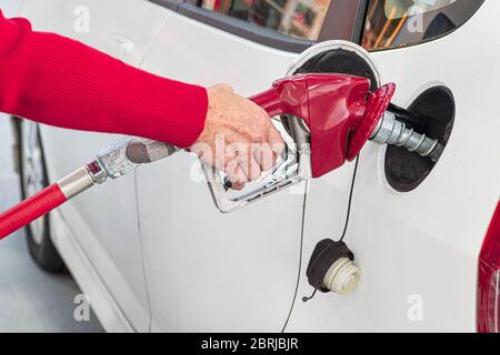 Horizontal close-up shot of an elderly woman’s hand pumping gasoline into her white car. Stock Photo