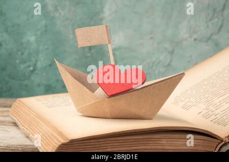Folded paper sailing boat and red heart on old opened book on table. Hand crafted origami paper sailing boat on an opened book Stock Photo