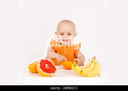 Happy baby sitting in high chair eating fruits in a white kitchen. Healthy nutrition for kids. Children eat oranges and grapefruits. Stock Photo