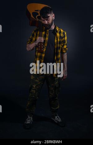 Tattooed man with a beard wearing a tartan yellow black shirt is  posing holding his guitar on his sholder Stock Photo