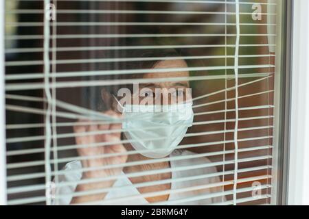 Worried elderly woman wearing face mask looking through window blinds during self-quarantine. Senior female wearing surgical mask. Stock Photo