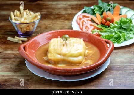 Portuguese francesine in a clay plate with salad and fried potatoes on a wooden background. Stock Photo