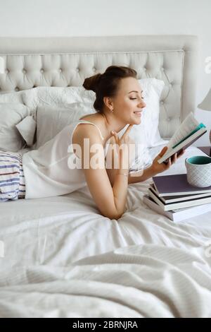 Happy young woomen relaxing on bed and reading book staying at home. Girl learning at home reading literature during quarantine Stock Photo