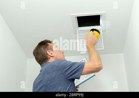 Man inspecting air ducts shining a flashlight through a small square ceiling vent into ducting pipes. Mature male examining the condition of air ducts Stock Photo