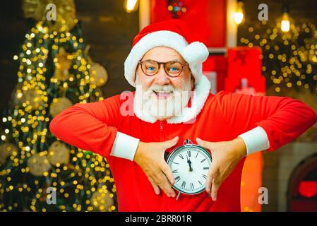 Thanksgiving day and Christmas. Santa Claus holding alarm clock against Christmas tree background. New year and Christmas time. Santa Claus at home Stock Photo