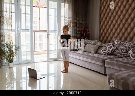 Cute little girl in sportswear watching online video on laptop and doing fitness exercises at home. Distant training with personal trainer, social Stock Photo